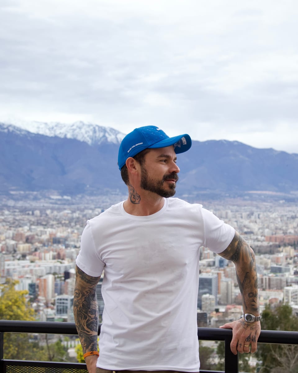 Davis Cabral de boné azul e camiseta branca, em um mirante com a cidade e a cordilheira ao fundo